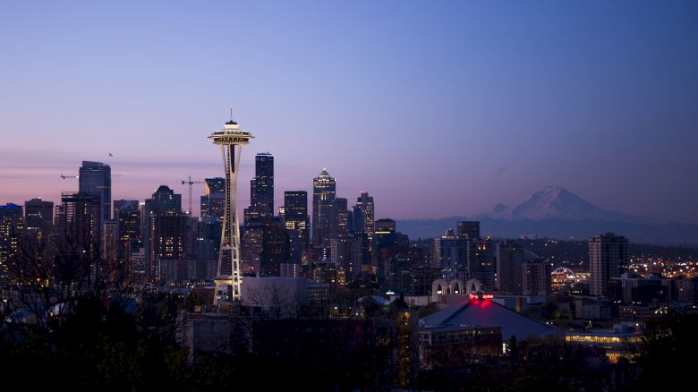 The image shows the Seattle skyline at twilight, featuring the iconic Space Needle with Mount Rainier in the background and city lights illuminating the scene.