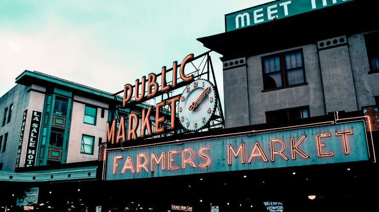 The image shows a neon sign for a public market and farmers market, with an iconic clock. Nearby buildings can be seen, including one labeled “The” hotel.