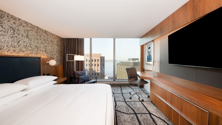 A modern hotel room with a large bed, a desk with a chair, a flat-screen TV, and a window view of buildings outside.