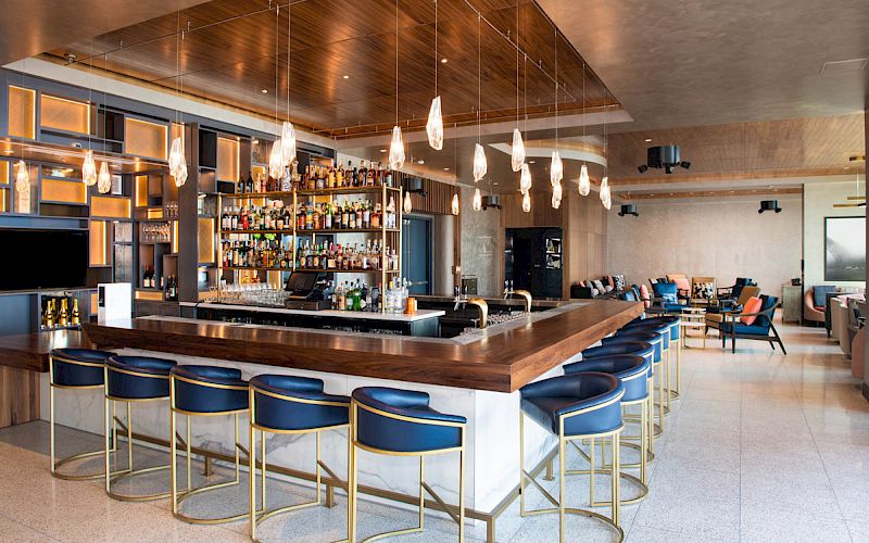 A modern bar with a wooden countertop, blue bar stools, and hanging lights, featuring a well-stocked liquor shelf and a comfortable seating area.