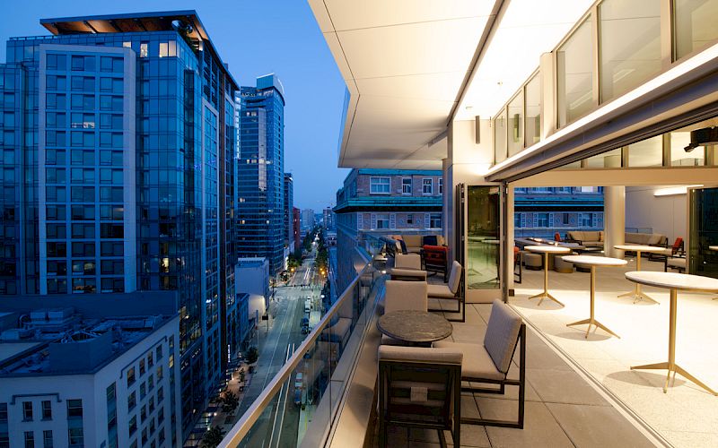 A modern balcony with outdoor seating overlooks a cityscape of tall buildings at dusk, with indoor seating visible through open doors.