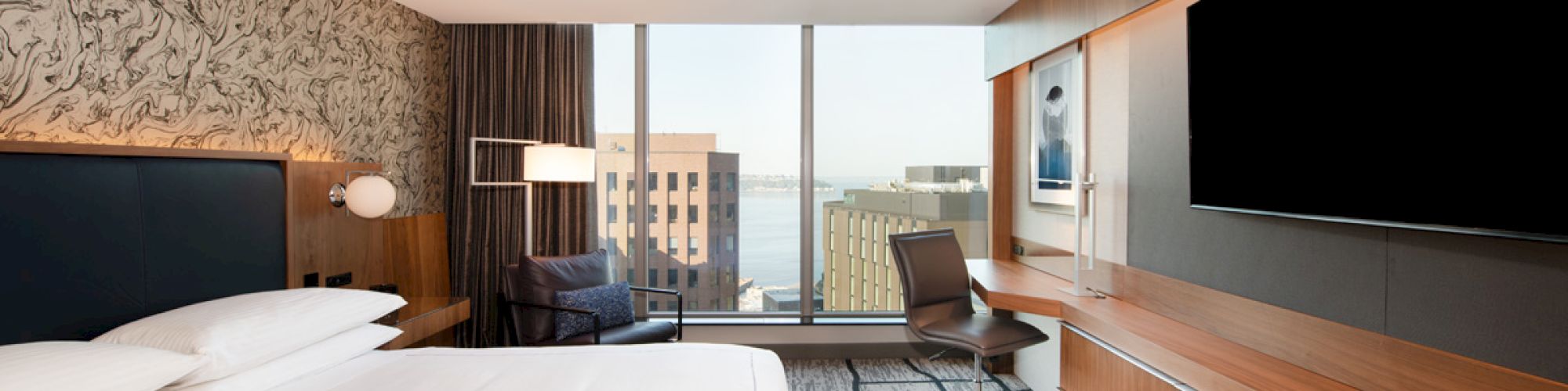 A modern hotel room features a large bed, a desk with a chair, a flat-screen TV, and a window with a city view.