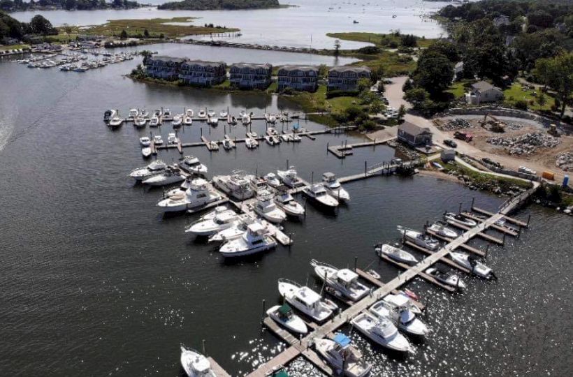 The image shows an aerial view of a marina with various boats docked, surrounded by water and a few buildings and trees along the shoreline.