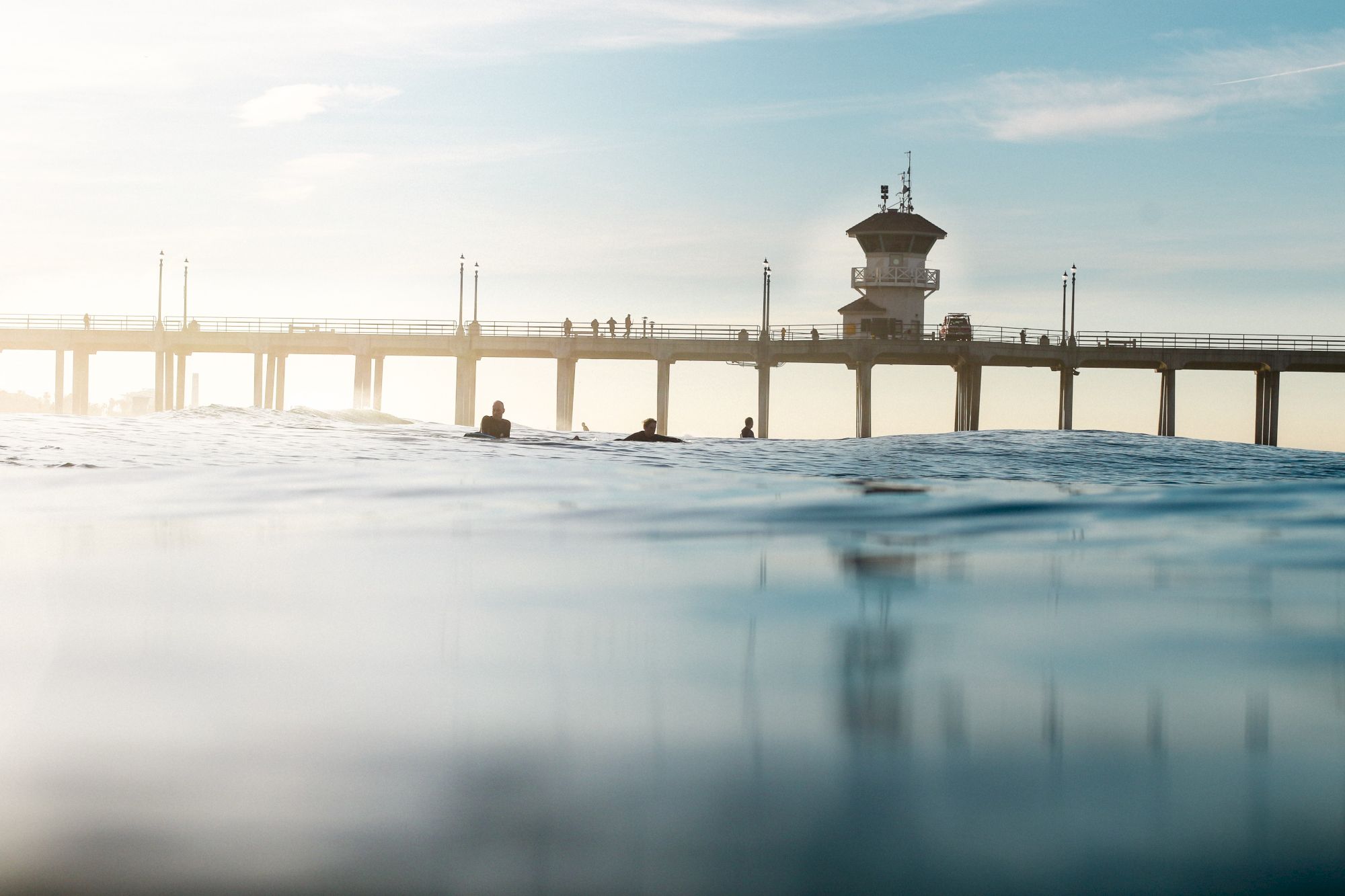 A pier extending over the ocean with a few surfers in the water and people walking on the pier under a clear sky during daylight.